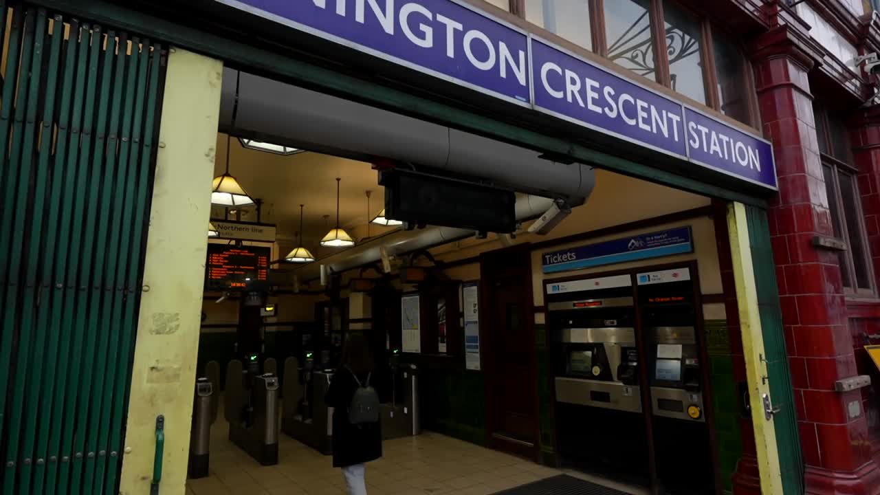Woman in casual attire taps card to enter Mornington Crescent Station at dusk. London, UK