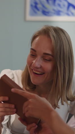 woman with disability in wheelchair gives partner book to check title on hardcover. blonde lady smiles sitting in living room with beloved girlfriend closeup