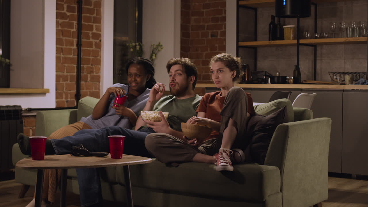 세 명의 룸메이트가 집에서 소파에 앉아 팝콘과 감자칩을 먹으면서 영화를 보고 있다 3