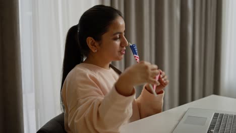 Side-view-of-a-happy-brunette-girl-in-a-beige-sweater-studying-foreign-languages-online-in-front-of-a-computer-at-home-and-showing-the-flag-of-the-United-States-of-America-to-the-webcam-in-a-modern-apartment-during-the-day