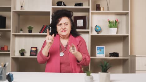 retrato de una mujer mayor sentada en una mesa regaña y agita sus manos, mirando a la cámara
