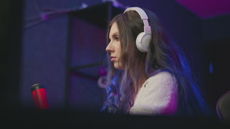 young female gamer wearing white headphones intensely focuses on computer screen while playing game in dark gaming room setup, with background slightly blurred and cool neon lighting in scene