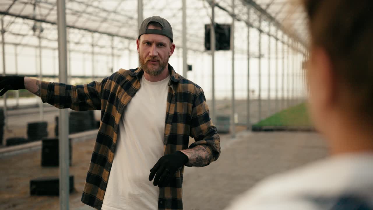 sobre su hombro un tipo confiado en una gorra con barba en una camisa a cuadros está hablando con una granjera y discutiendo planes para trabajar en un invernadero en la granja