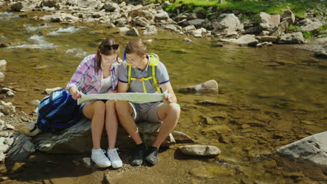 A-Young-Couple-Is-Sitting-On-A-Rock-Near-A-Mountain-River-They-Look-At-The-Map-Together-Planning-The