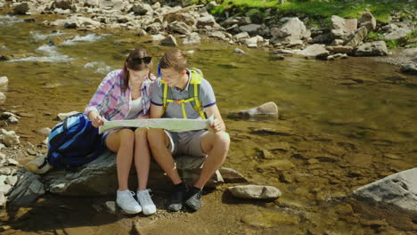 a young couple is sitting on a rock near a mountain river they look at the map together planning the