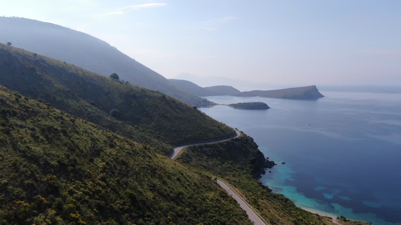Drone view in Albania flying over a green hill, a narrow road and a blue ocean on a sunny day