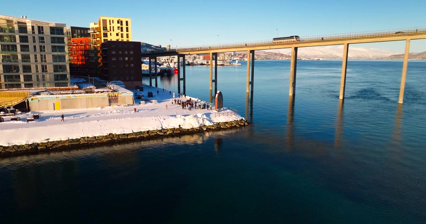 Riva coperta di neve del moderno quartiere di Vervet e Ponte di Tromso sull'acqua blu del fiordo in inverno, Norvegia. Vista aerea laterale da drone, spazio per il testo.
