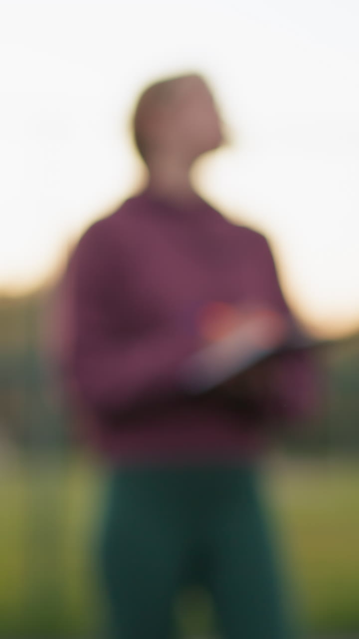 Close-up of volleyball held by athlete in green sweater, preparing to serve, with blurred background featuring coach observing and people walking across field in outdoor training session