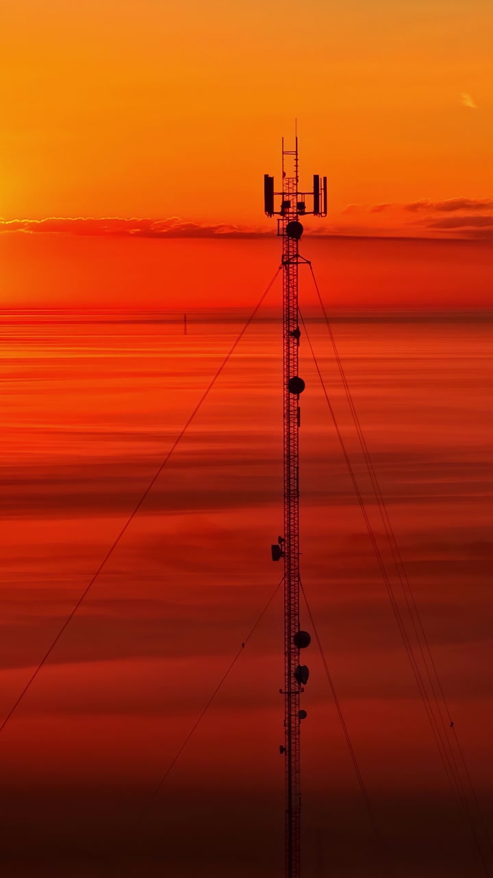 Vista aerea verticale, torre di telecomunicazioni in Lettonia stagliata contro un vivido cielo arancione al tramonto, a simboleggiare la tecnologia wireless, la connessione e l'infrastruttura rurale