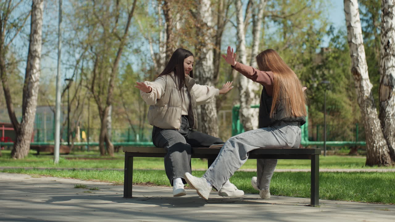 Девушки на скамейке в парке, азиатка и европейка, энергично хлопают в ладоши, радостно приветствуя друг друга, солнечный весенний день в березовой роще, непосредственная юношеская радость близких подруг, игривые движения
