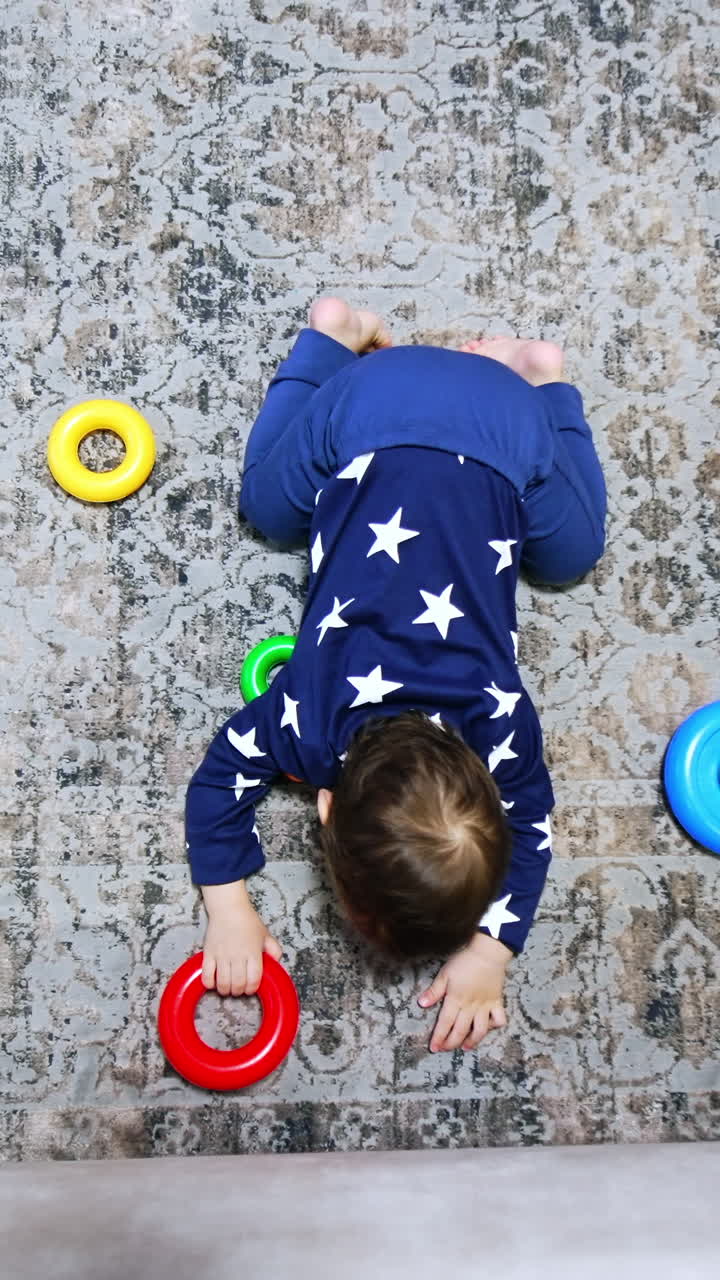 Little baby boy lies kneeling on the floor of the room. Toy rings are scattered around. Kid raises his head looking at camera. Top view. Vertical video