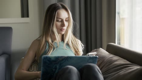 mujer rubia sentada en el sofá con una computadora portátil en las rodillas usando su teléfono celular