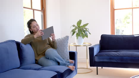 Happy-biracial-woman-sitting-on-sofa,-smiling-and-using-tablet-in-sunny-living-room