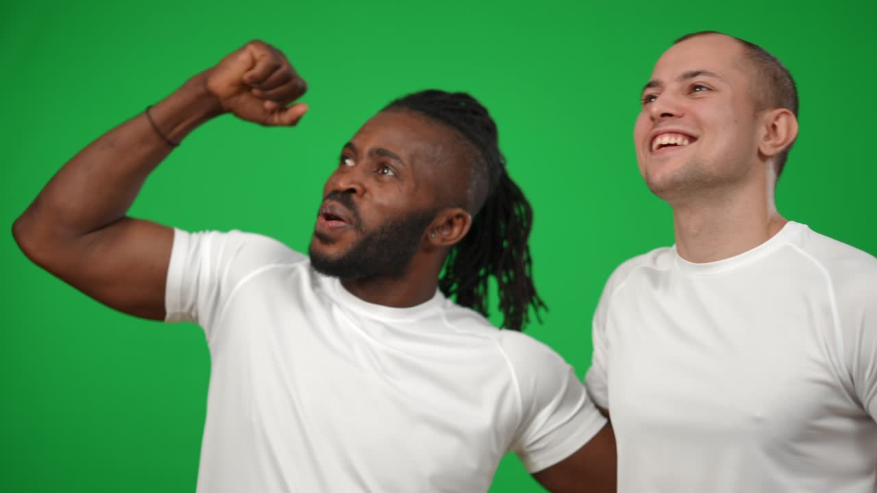 Joyful Caucasian and African American sportsmen hugging looking away gesturing high-five at green screen. Happy professional footballers cheering rejoicing success at chromakey background.