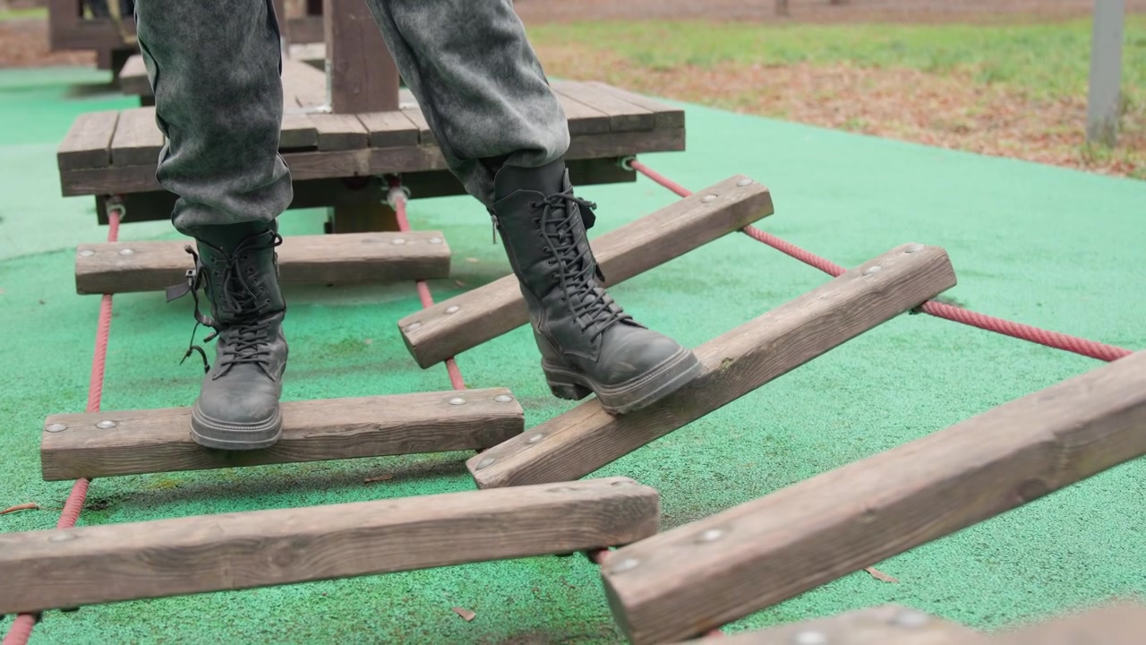 Boots stepping across wooden planks on suspended ladder, laced combat boots placing careful foot after foot, green safety surface and taut ropes visible, slow focused balance and steady motion
