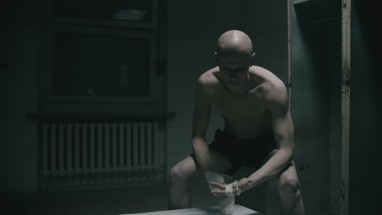 Bleik, skallet, tynn mann bandasjerer hendene sine på det skumle gamle badet om natten.