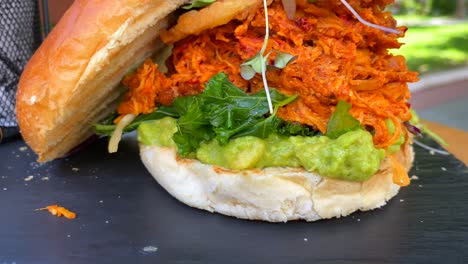 Juicy-pulled-chicken-burger-with-onion-rings,-avocado,-crispy-kale-and-a-brioche-bun,-fast-food-restaurant-with-nature-view,-4K-shot