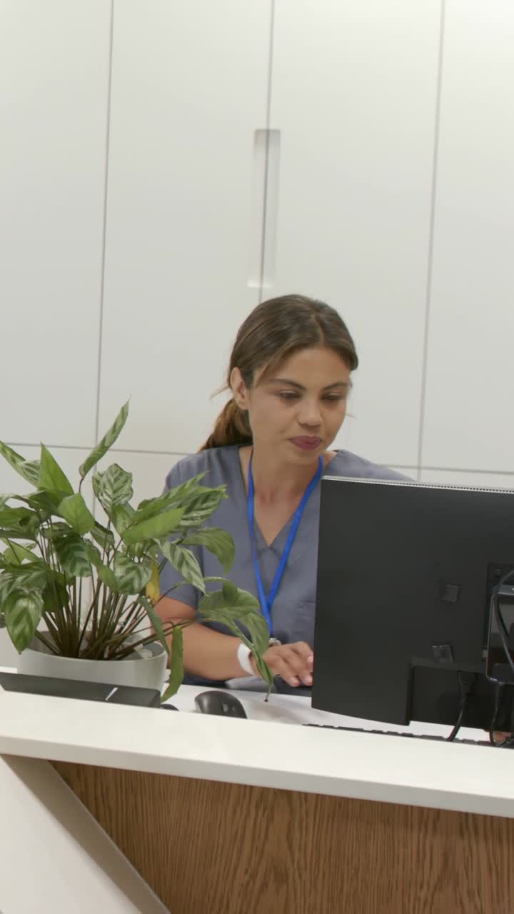 Vertikální video: Žena v uniformě se letmo podívá doleva, natahuje se pro myš a kliká, zatímco pracuje na recepci.