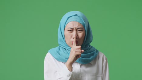 close up of asian muslim woman looking at camera and making shh gesture while standing in the green screen background studio