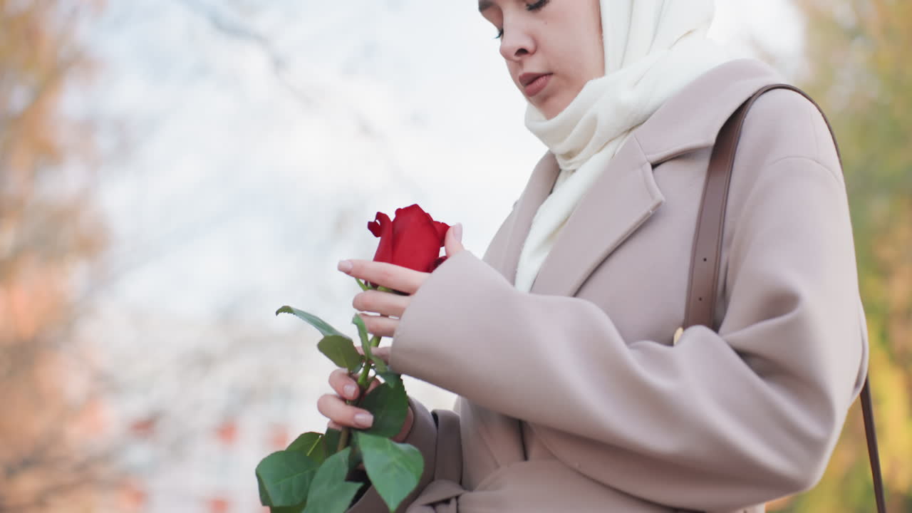 Seorang wanita muda Kaukasia menemukan mawar merah, bangku taman kosong, daun-daun musim gugur, mengenakan mantel krem dan syal putih, mengangkat tangkai dengan lembut, dengan ekspresi merenung, menciptakan suasana kerinduan dan kenangan di bawah cahaya mendung yang lembut.