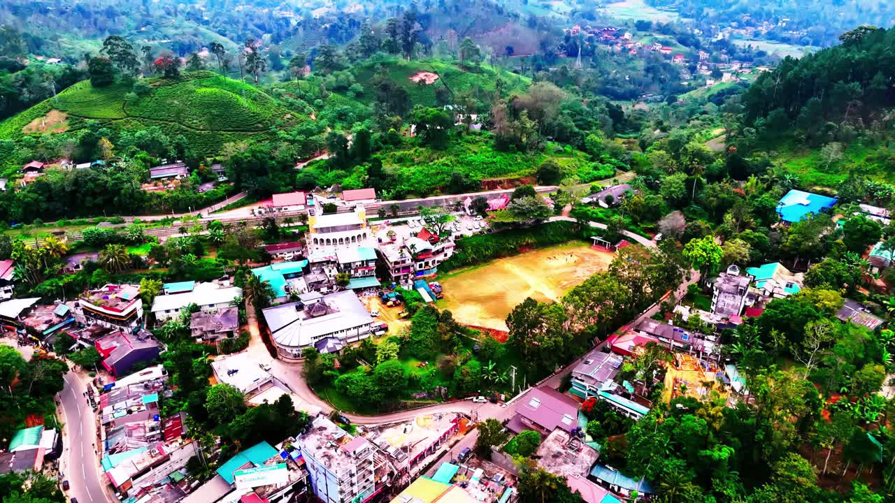 A lively hillside village in Sri Lanka features winding roads, colorful rooftops, and a central field, all surrounded by lush green tea plantations and forested hills stretching into the distance.