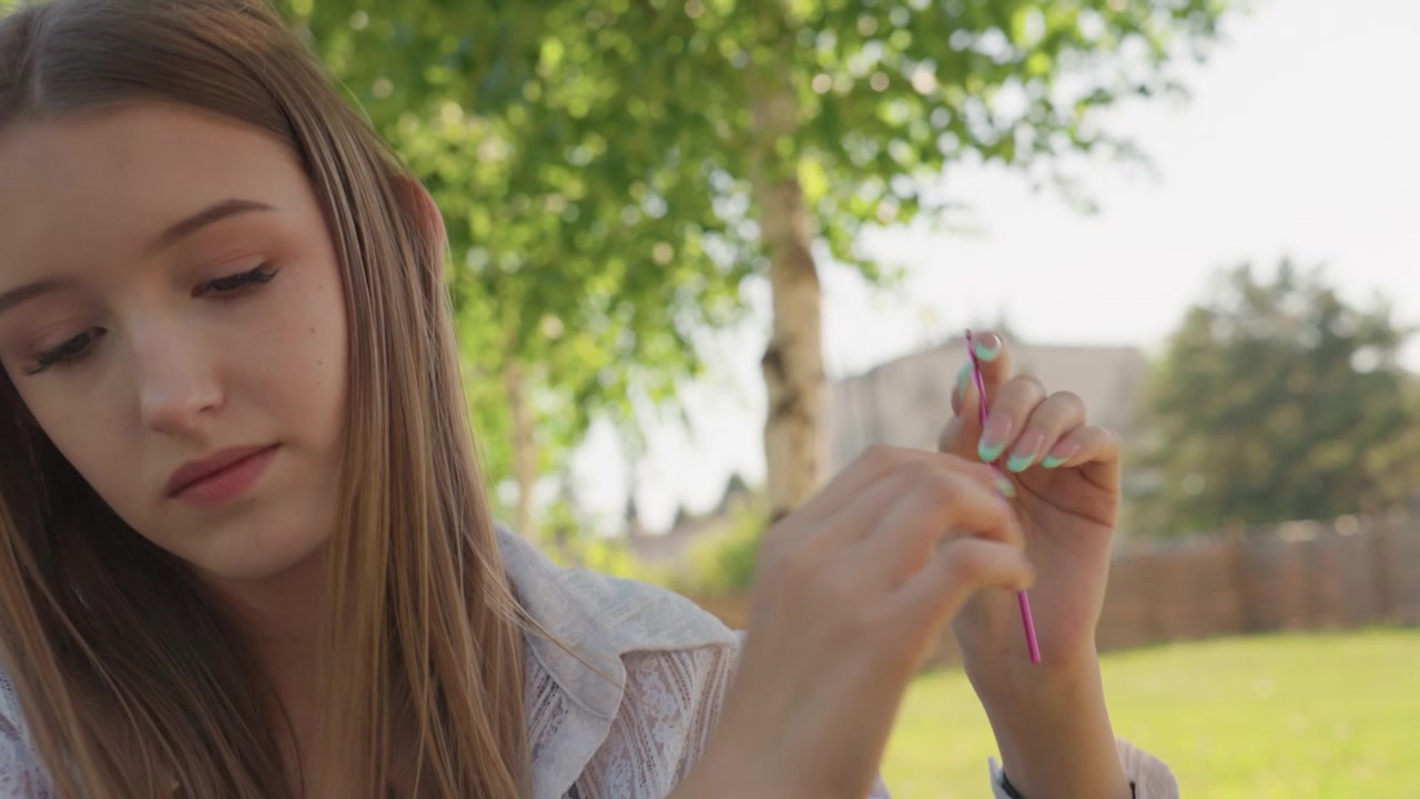 Close up of young woman sitting outdoors on bench holding and examining crochet hooks from colorful packaging. Woman wears light colored lace fabric over dark top with mint green nail polish