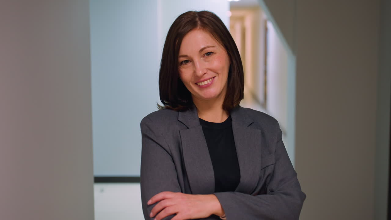 Professional woman in business attire standing confidently with arms crossed in modern office hallway. Serious expression with neutral background suggesting leadership, business