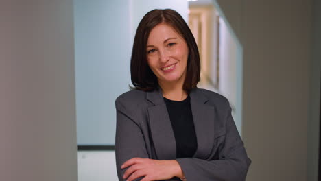 professional woman in business attire standing confidently with arms crossed in modern office hallway. serious expression with neutral background suggesting leadership, business