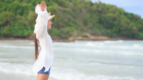 Close-up-of-a-young-woman-in-shorts,-white-cover-up,-and-sun-hat-joyfully-walking-into-the-surf