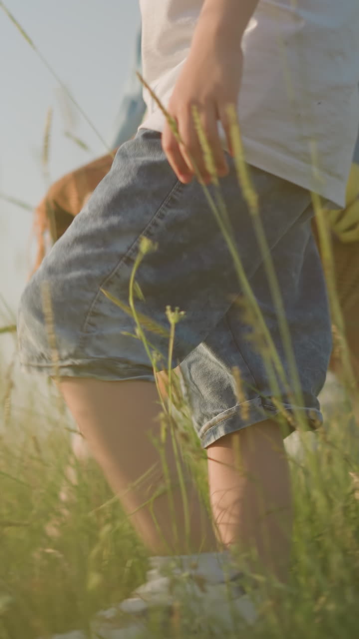 A close-up of a young boy in denim shorts walking alongside a woman in a blue dress holding a basket of fruit, as they stroll through a sunlit grassy field
