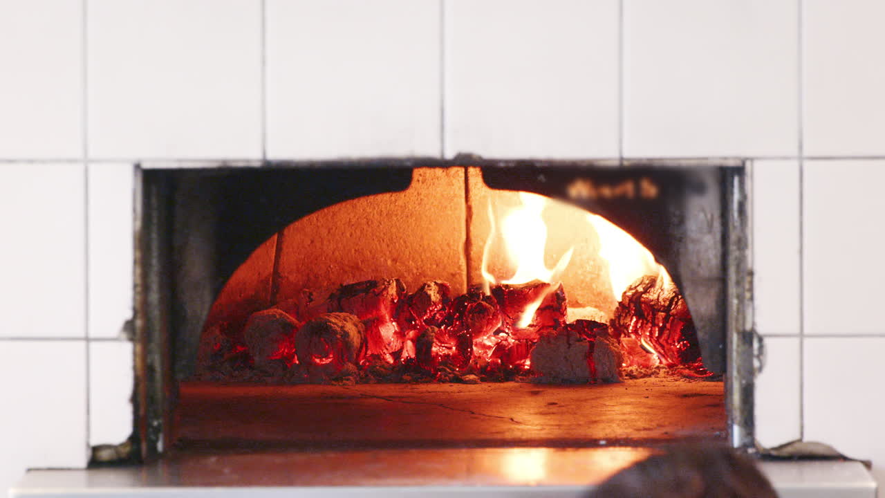 한 남자가 목공의 집에서 점토 오븐에서 불타는 뜨거운 석탄에 불타는 나무를 추가합니다.