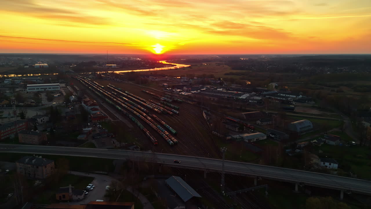 Stunning aerial view captures a vibrant sunset over the Jelgava train station in Latvia with many freight trains on tracks and a river in the distance