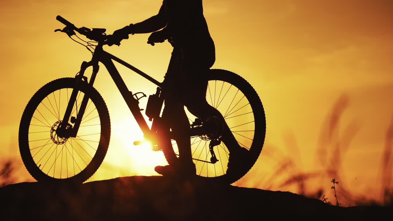 Sportsman biker walk with bike outdoors at golden sunset. Silhouette of a bicycle and a cyclist walking in the evening against the beautiful summer sunset. Close-up.