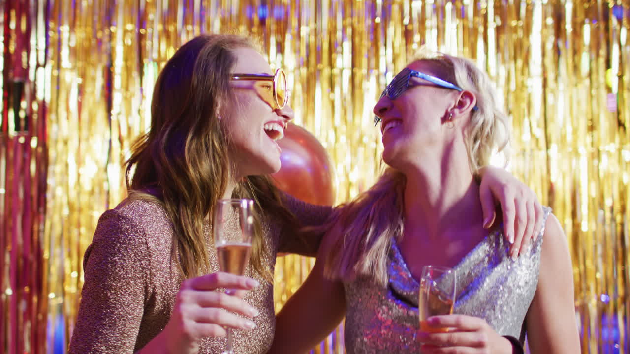 Video of two happy caucasian female friends dancing with glasses of champagne at a nightclub