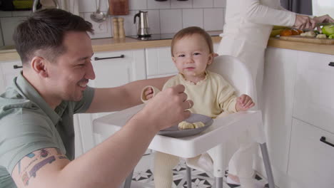 happy father feeding his cute baby girl sitting in her high chair in the kitchen while mother cutting fruit behind them