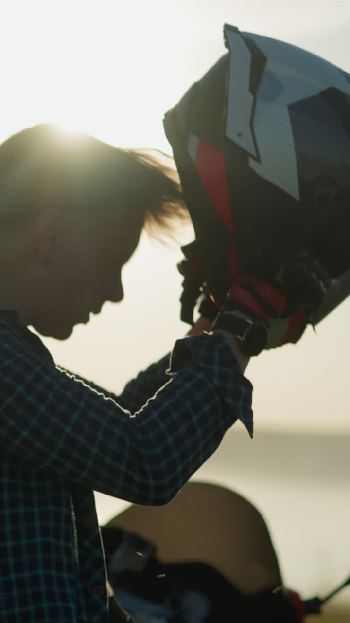 una mujer joven, en un campo cubierto de hierba, se quita el casco y ajusta su cabello mientras la brillante luz del sol se refleja en su figura, se para junto a una bicicleta