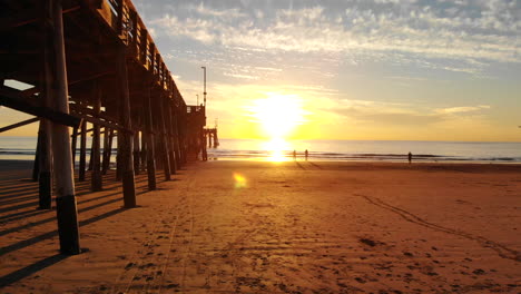 drone flying under newport beach pier at sunset as sunlight beams and people walk the california coast in silhouette aerial