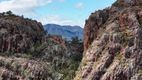 drone camera smoothly pans across rugged split rock cliffs, revealing dramatic rock formations, native vegetation, and distant mountain ranges under bright daylight