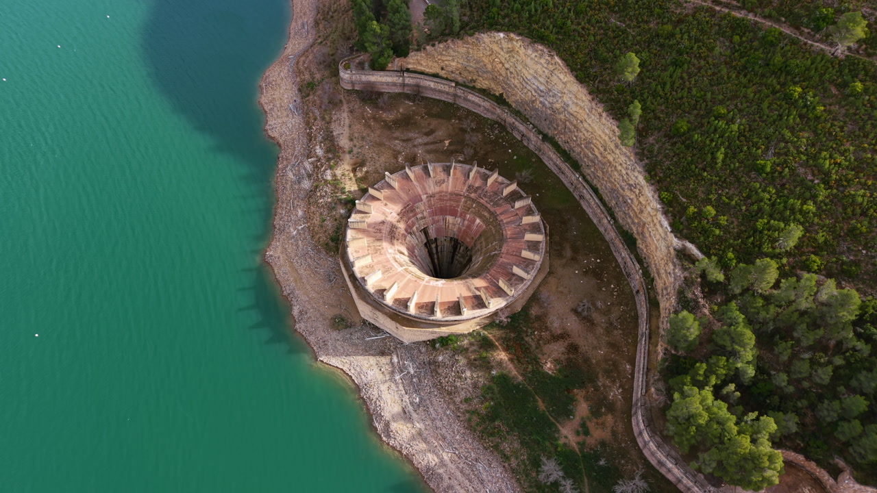 Overhead view of a circular concrete intake tower at Benageber gravity dam beside a turquoise reservoir in Valencia, Spain