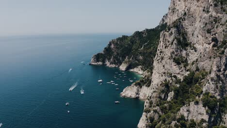 Drone-shot-of-Capri,-Italy's-steep-cliffs-leading-to-boat-filled-warm-water