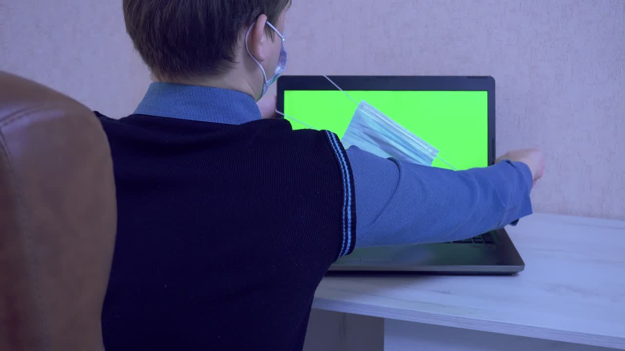 un tipo con una máscara médica se pone una máscara protectora en una computadora portátil con un interlocutor virtual con el que se comunica a través de enlace de video como un símbolo de protección contra un virus, pantalla verde