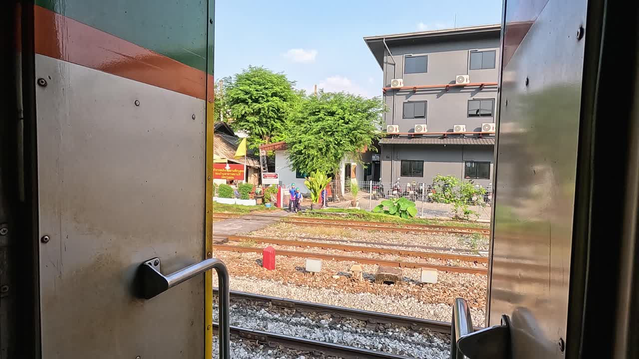 Daytime view from open train doorway passing Bangkok railway yard, tracks, apartments, and greenery
