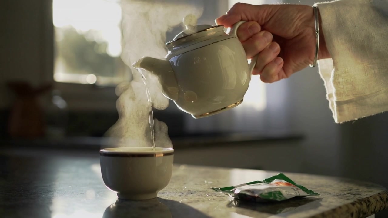 Una mano ammanettata con un braccialetto inclina una teiera versando il tè in una tazza sul bancone in cucina, bustina.