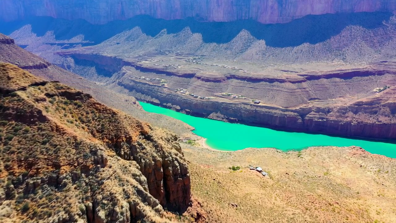 La telecamera si muove fluidamente lungo il bordo di un canyon arido, mantenendo l'acqua turchese brillante centrata in basso mentre scogliere texturizzate, vegetazione rada e formazioni rocciose stratificate si spostano attraverso l'inquadratura