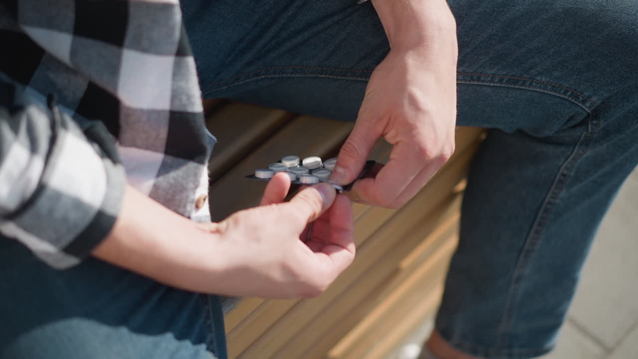 Close up of boy wearing jeans and plaid shirt pressing tablet from blister pack while seated on wooden bench in sunny outdoor setting with warm lighting and relaxed posture
