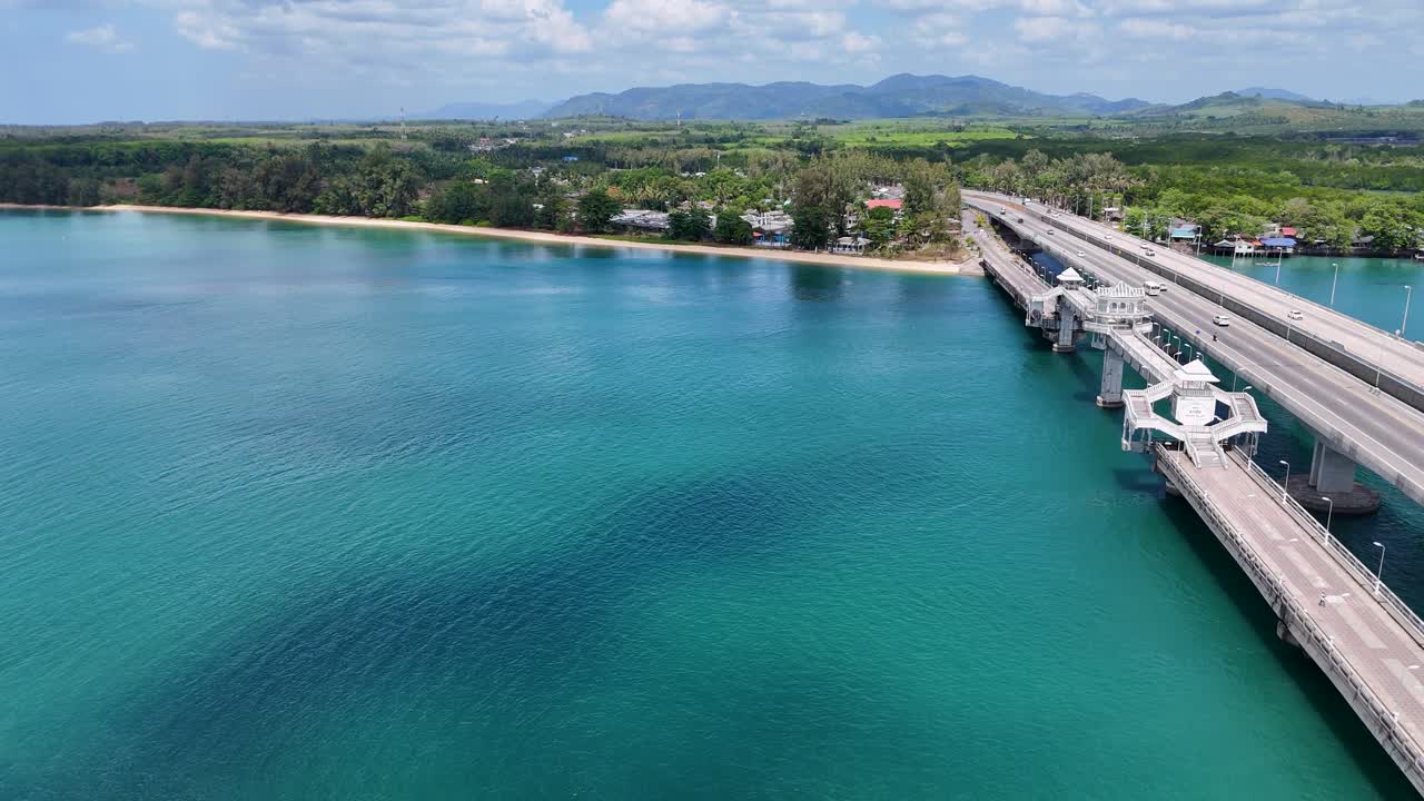 Drone footage captures Sarasin Bridge in Phuket, Thailand, spanning turquoise waters under clear skies, highlighting the scenic coastal landscape