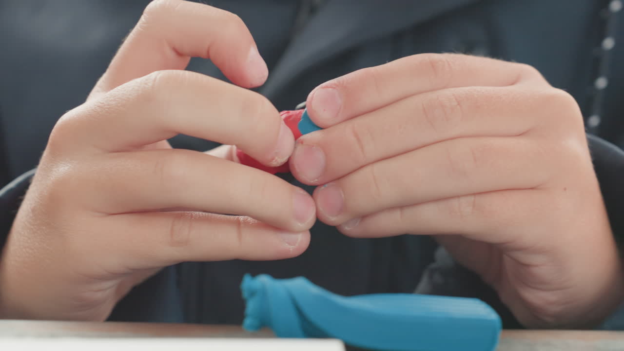 Primo piano delle mani di un bambino caucasico che modellano l'argilla, pasta modellabile rossa e blu sul tavolo, dito concentrato che preme, giacca testurizzata visibile, esercizio di apprendimento tattile, calma concentrazione, preparazione alla scuola.