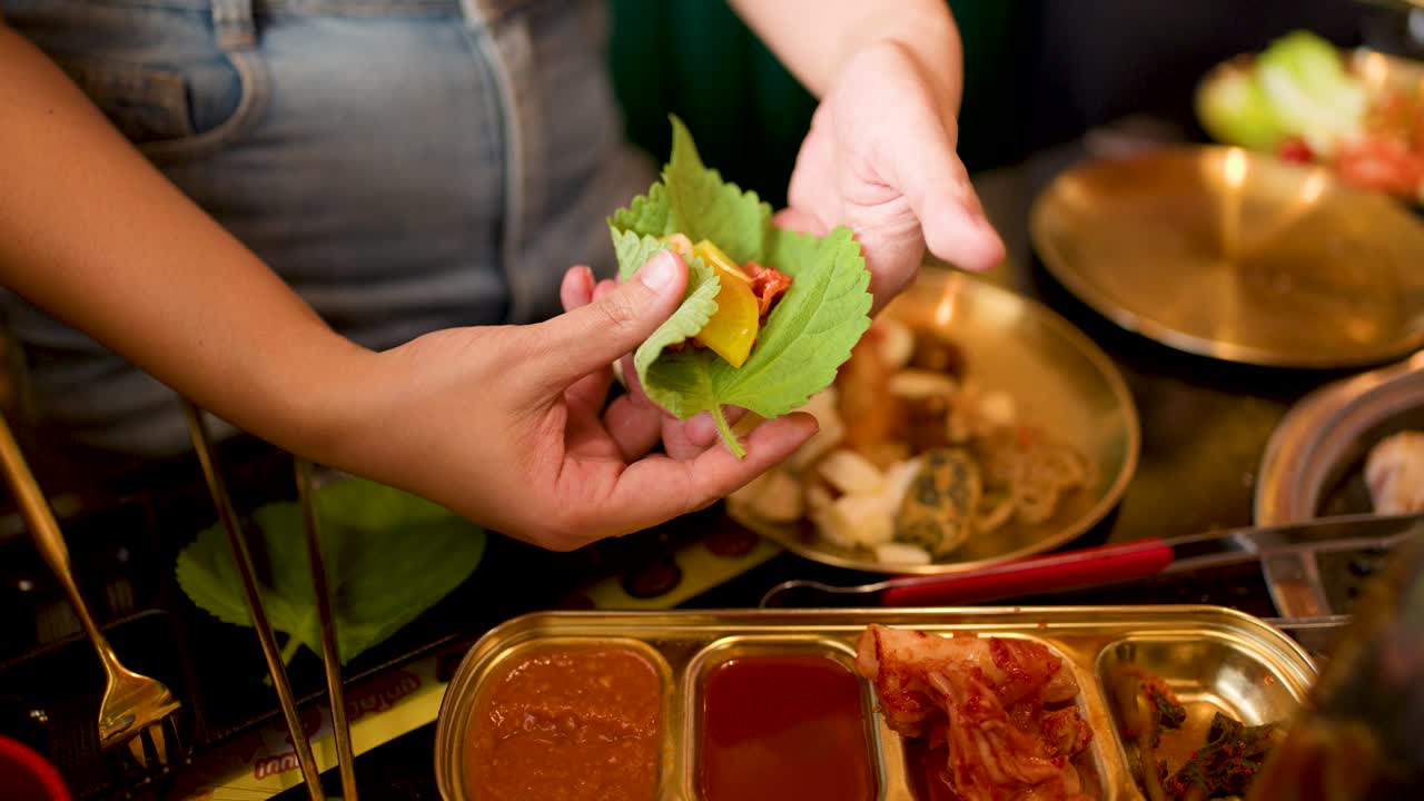 Person prepares Korean lettuce wrap with pork, kimchi, dipping sauces at restaurant table, warm lighting