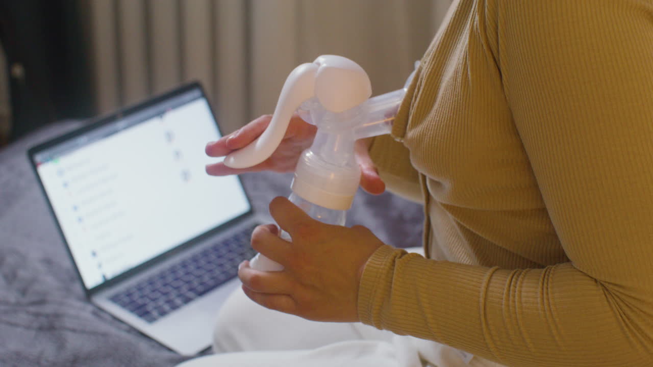 Close Up Of A Woman Using Breast Pump While Sitting On The Bed With Laptop Computer At Home