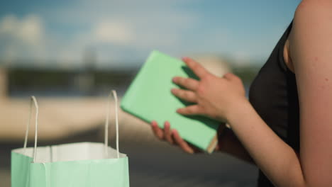 close-up of woman seated on outdoor bench with shopping bags on both sides, she retrieves a book from a mint green bag, opens it, and flips through the pages, with blurred background